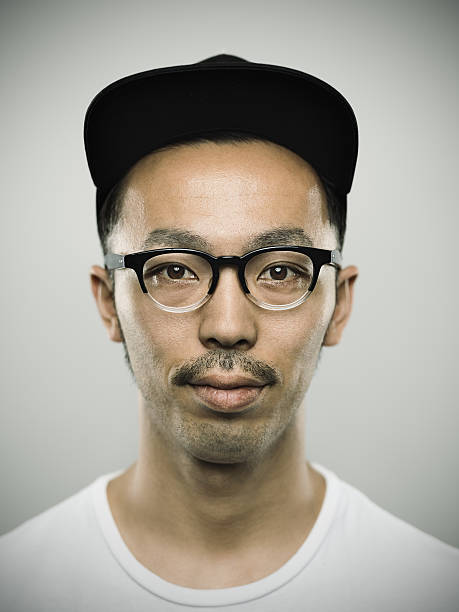 Portrait of a young japanese man looking at camera Studio portrait of a japanese young man looking at camera with relaxed expression. The man has around 25 years and has short hair and casual clothes, wearing a baseball cap and glasses. Vertical color image from a medium format digital camera. Sharp focus on eyes. thin moustaches stock pictures, royalty-free photos & images