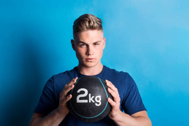 Portrait of a young man with heavy ball in a studio. Portrait of a young man holding heavy medicine ball in a studio, standing on a blue background. Copy space. teenage boys men blond hair muscular build stock pictures, royalty-free photos & images