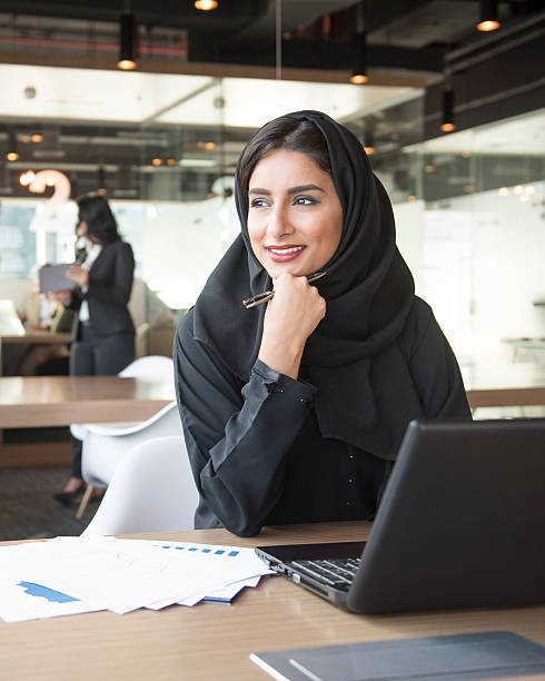 Portrait of a young Middle Eastern businesswoman at work Attractive young ethnic Arab businesswoman in traditional clothing using laptop, smiling and looking away. Young Arabic woman with hand on chin holding pen. middle eastern woman stock pictures, royalty-free photos & images
