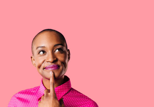 Portrait of a young woman in pink shirt with finger on mouth looking up thinking, isolated on pink studio background stock photo