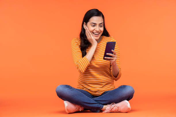 portrait of a young women using mobile phone sitting isolated over orange background stock photo