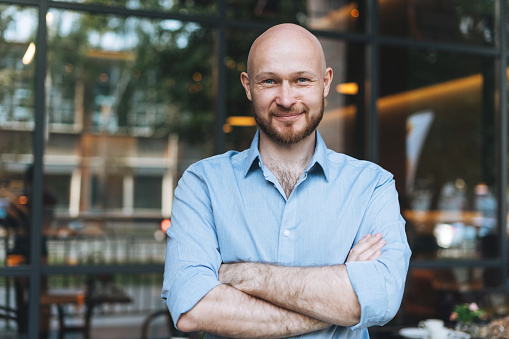 Portrait of Adult bald smiling attractive man forty years with beard in blue shirt businessman against glass wall of street cafe
