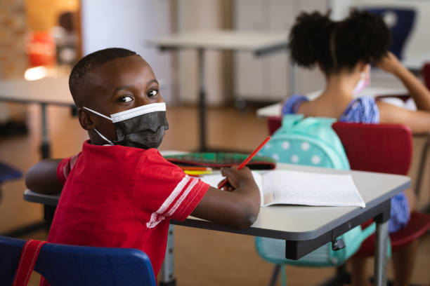 Portrait of african american boy wearing face mask sitting on his desk in class at elementary school stock photo