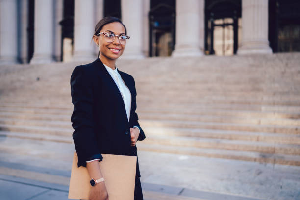 Portrait of African American businesswoman with successful career dressed in formal wear standing against architecture office building with folder in hands. Experienced woman entrepreneur smiling stock photo