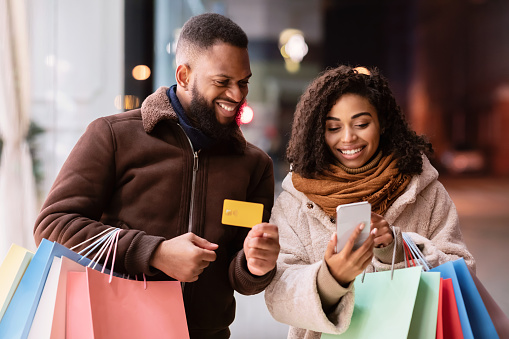 Ordering Online. Smiling black casual couple using cellphone, man showing debit credit card, family holding colorful shopping bags, walking outdoors in the evening, making purchases on internet