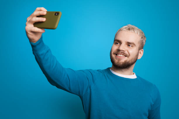 Portrait of an caucasian man taking a selfie while standing and looking at the screen of his phone isolated over blue background. stock photo