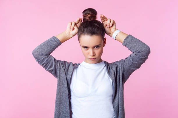 Portrait of angry brunette teenage girl making bull horn gesture with fingers on head. isolated on pink background Portrait of angry brunette teenage girl with bun hairstyle in casual clothes making bull horn gesture with fingers on head, looking bully and hostile. indoor studio shot isolated on pink background ugly girl stock pictures, royalty-free photos & images