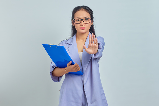 Portrait of angry young Asian woman holding folder and says no shows prohibition gesture refuses something isolated on purple background