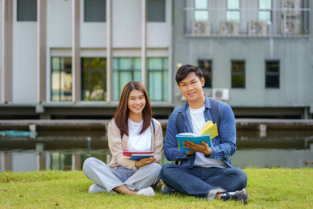 Portrait of Asian man and woman university student sitting on grass in campus looking at camera and smile and reading a book together in park. stock photo