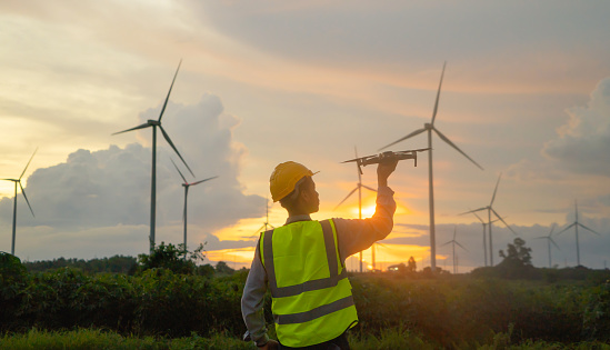 Portrait of Asian windmill engineer man, worker working, control a drone on site at wind turbines field or farm, clean energy source. Eco technology for electric. industry nature environment. People
