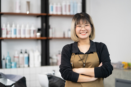 Portrait of Asian women hair stylish business owner standing and smile inside of hair salon with shampoo and hair shower area as the background. Beauty and fashion, personal care business.