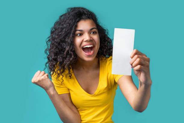 Portrait of attractive young african american girl Close up cropped portrait of young african american woman holding blank lottery ticket or coupon in hand and clenching her fist making winning gesture, screaming "yes" celebrating success. lottery stock pictures, royalty-free photos & images