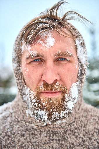 Portrait of bearded man with snow on his face. Man is frozen in pullover with hood and flying hair.