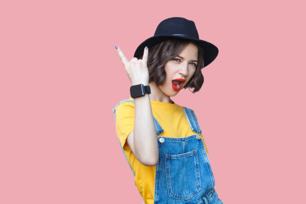 Portrait of beautiful amazed young woman in yellow t-shirt, blue denim overalls with makeup and black hat standing with rock horns, screaming and looking at camera Portrait of beautiful amazed young woman in yellow t-shirt, blue denim overalls with makeup and black hat standing with rock horns, screaming and looking at camera. studio shot on pink background. rock musician stock pictures, royalty-free photos & images