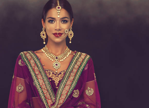 Portrait of beautiful girl dressed in a traditional indian national suit, jewelry set, blouse and shawl (dupatta). Young attractive woman, dressed in a traditional indian suit-sari, with purple blouse and shawl (dupatta) with gilded hand-made decoration. Posh jewelry set is consisting of big earrings, bright necklace, head adornment (tikka) and gold nose ring. indian jewelry stock pictures, royalty-free photos & images