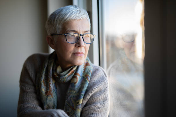 Portrait of beautiful mature woman relaxing by the window Smiling senior woman day dreaming and looking through the window during her coffee time. looking through window stock pictures, royalty-free photos & images