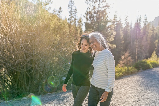 A beautiful mixed race young adult woman embraces her vibrant retirement age mother. The mother and daughter are enjoying a relaxing walk in nature on a beautiful, sunny day. In the background is a mountainous evergreen forest bathed in sunlight.