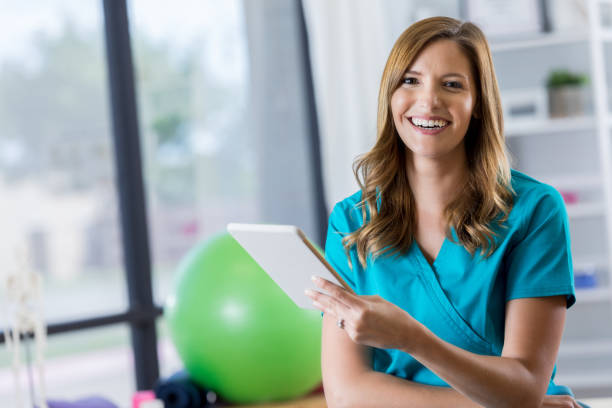 Portrait of beautiful physical therapist Beautiful mid adult Caucasian physical therapist holds a digital tablet. The tablet contains a patient's treatment plan. She is smiling cheerfully at the camera. yoga ball work stock pictures, royalty-free photos & images