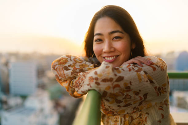 Portrait of beautiful woman looking at camera and smiling at balcony during sunset stock photo