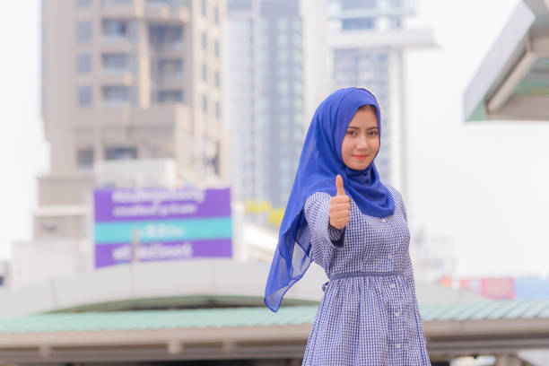 Portrait of beautiful young muslim woman in hijab dress standing smiling and thumb up on outside in city. stock photo