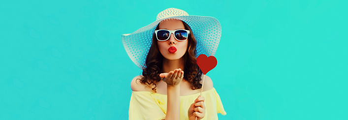 Portrait of beautiful young woman blowing her lips with lipstick with red sweet heart shaped lollipop wearing a summer straw hat on blue background