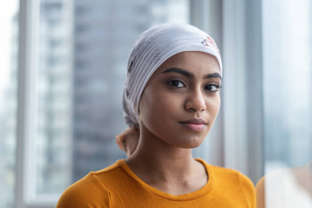 Portrait of beautiful young woman with cancer A courageous Malaysian woman with cancer wears a bandana to hide her hair loss. She is leaning against a window and is looking directly at the camera. She has a thoughtful and serious expression on her face. survival stock pictures, royalty-free photos & images