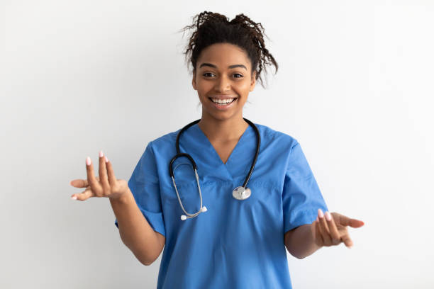 Portrait of black doctor smiling and talking to camera Professional Specialist. Smiling black female doctor wearing blue medical uniform coat and stethoscope talking to camera, explaining and gesturing, standing isolated over white studio background wall nurse talking to camera stock pictures, royalty-free photos & images