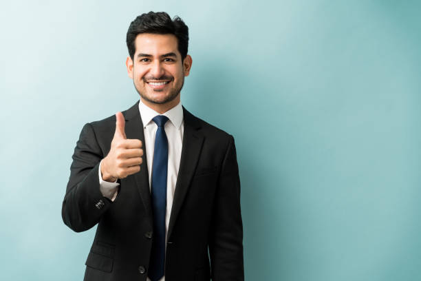 Portrait Of Businessman Showing Hand Sign In Studio Smiling Hispanic male executive gesturing thumbs up against isolated background business thumbs up stock pictures, royalty-free photos & images