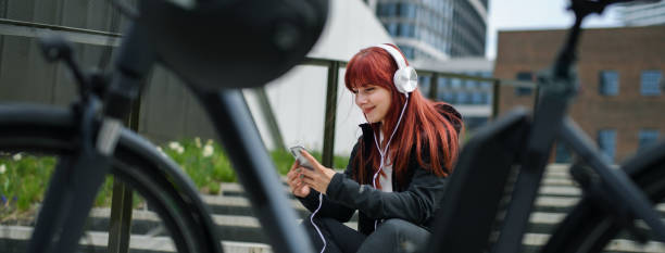 Portrait of businesswoman commuter on the way to work with bike, resting listening to music, sustainable lifestyle concept. Wide shot. stock photo