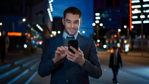 het portret van kaukasische zakenman in een kostuum gebruikt een smartphone op donkere straat in de avond. hij ziet er zelfverzekerd en succesvol uit. atmosferische stedelijke lichten van de stad op de achtergrond. - pakjesavond stockfoto's en -beelden