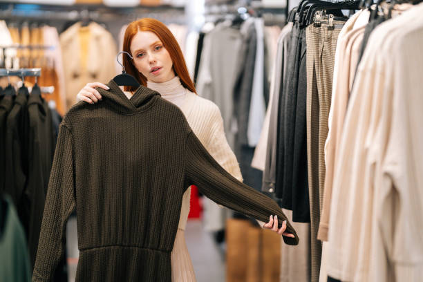 Portrait of charming young woman choosing clothes and looking to mirror in clothing store. stock photo