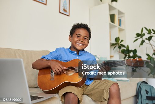istock Portrait of cheerful adolescent black boy sitting on sofa in living room and playing guitar according to video lesson 1264349000