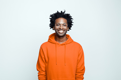 Happy afro american young man wearing orange hoodie and glasses, smiling at camera. Studio shot on grey background.