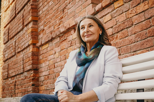 Portrait of cheerful trendy looking gray haired retired woman sitting on bench outdoors against red brick wall wearing elegant white jacket, having dreamy thoughtful joyful facial expression
