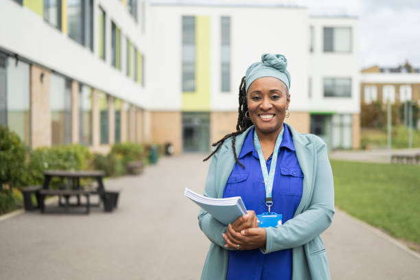 Portrait of confident Black female educator on campus Waist-up front view of mature woman in professional attire with headscarf and ID lanyard standing outdoors holding composition booklets and smiling at camera. school counselor stock pictures, royalty-free photos & images