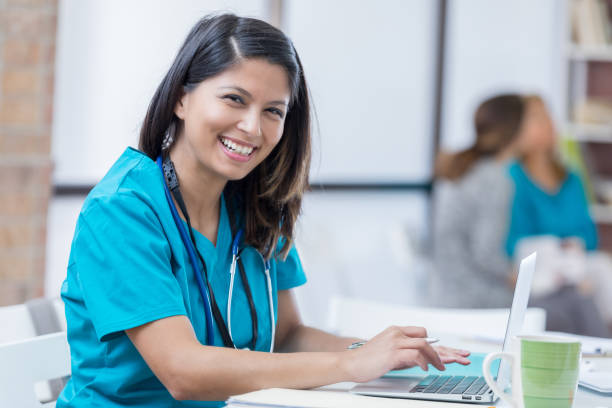 Portrait of confident healthcare professional Beautiful mid adult female Filipino nurse or doctor uses a laptop in her office. She is wearing scrubs and a stethoscope. She is smiling at the camera. filipino woman stock pictures, royalty-free photos & images