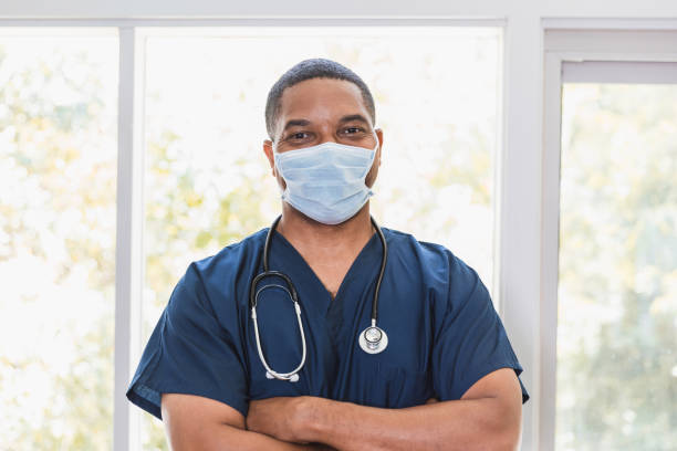 Portrait of confident male home healthcare nurse A confident African American male home healthcare nurse looks at the camera while standing with his arms crossed. He is wearing a protective face mask. males stock pictures, royalty-free photos & images