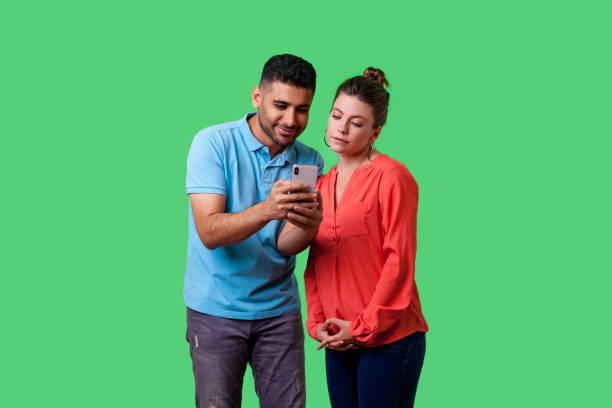 Portrait of curious attractive young couple in casual wear using mobile phone. isolated on green background, indoor studio shot Portrait of curious attractive young couple in casual wear standing, reading message together on mobile phone, looking concentrated and interested. isolated on green background, indoor studio shot smart phone green background stock pictures, royalty-free photos & images