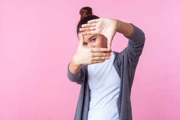 verticale d'adolescente brune curieuse regardant par le geste de cadre de photo fait des mains. isolé sur le fond rose - focus photos et images de collection