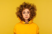 Portrait of cute curly-haired woman in urban style hoodie standing with puffed cheeks, holding breath and looking at camera with big eyes, amazed expression. indoor studio shot, yellow background