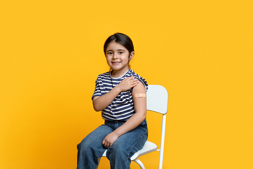 Portrait Of Cute Little Arab Girl Showing Arm After Coronavirus Vaccination, Adorable Middle Eastern Child Got Covid-19 Vaccine Injection, Sitting With Rolled Up Sleeve Over Yellow Background