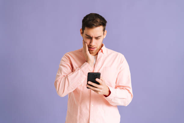 Portrait of disappointedyoung man holding phone with doubtful and skeptical expression on pink isolated background. stock photo