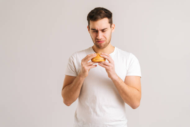 Portrait of dissatisfied young man with disgust eating bad burger on white isolated background. stock photo