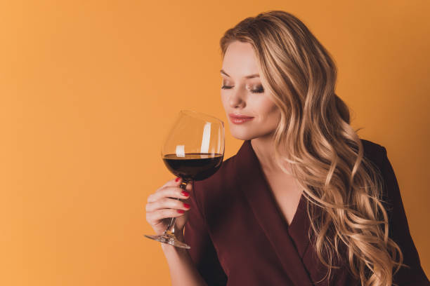 portrait of dreamy lovely cute woman with close eyes holding glass with red wine in hand enjoying smell of beverage having fun isolated on dark yellow background - sniffing glass imagens e fotografias de stock