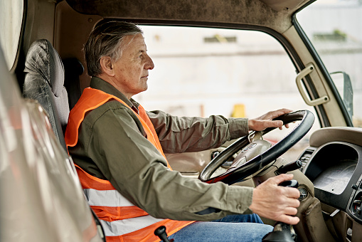 Side view of Caucasian man wearing casual clothing and reflective vest, looking straight ahead with one hand on steering wheel and other on gearshift.