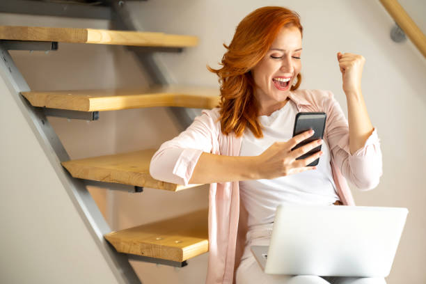Portrait of euphoric young woman holding phone reading good news, while sitting over staircase at home. stock photo