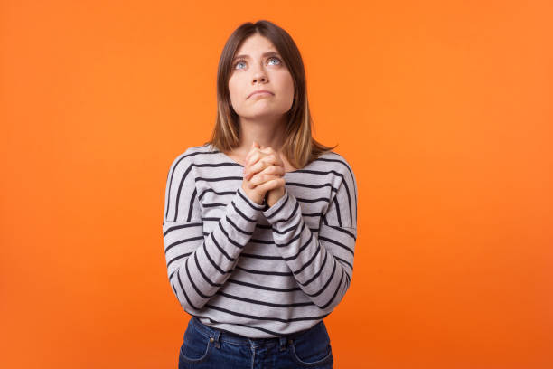 Portrait of faithful young woman with brown hair in long sleeve shirt. indoor studio shot isolated on orange background Portrait of faithful young woman with brown hair in long sleeve shirt standing, looking up and holding hands in prayer, pleading help or apology. indoor studio shot isolated on orange background prayer request stock pictures, royalty-free photos & images