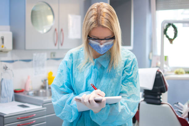 Portrait of female dentist in a protective suit, shield, mask and eyeglasses writing notes to planner book in dental office. stock photo