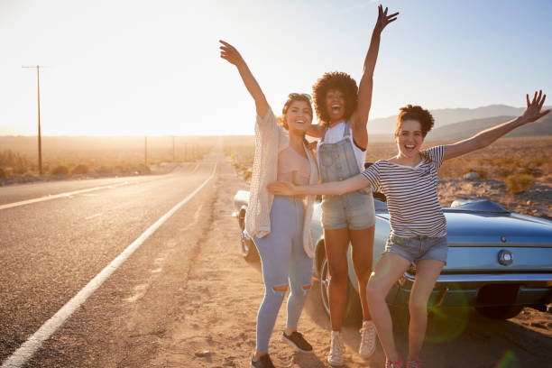 Portrait Of Female Friends Enjoying Road Trip Standing Next To Classic Car On Desert Highway Portrait Of Female Friends Enjoying Road Trip Standing Next To Classic Car On Desert Highway girlfriend stock pictures, royalty-free photos & images