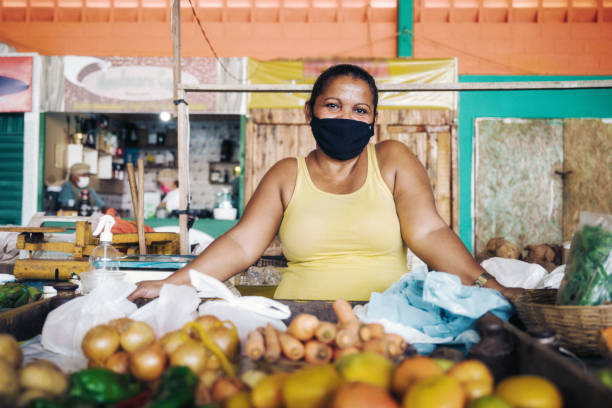 portrait of female market vendor with face mask behind her vegetable stall at public market stock photo
